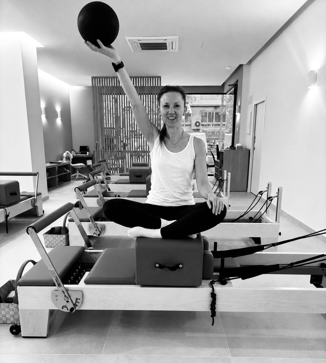 Maud sitting cross-legged on a reformer, holding a ball above her head